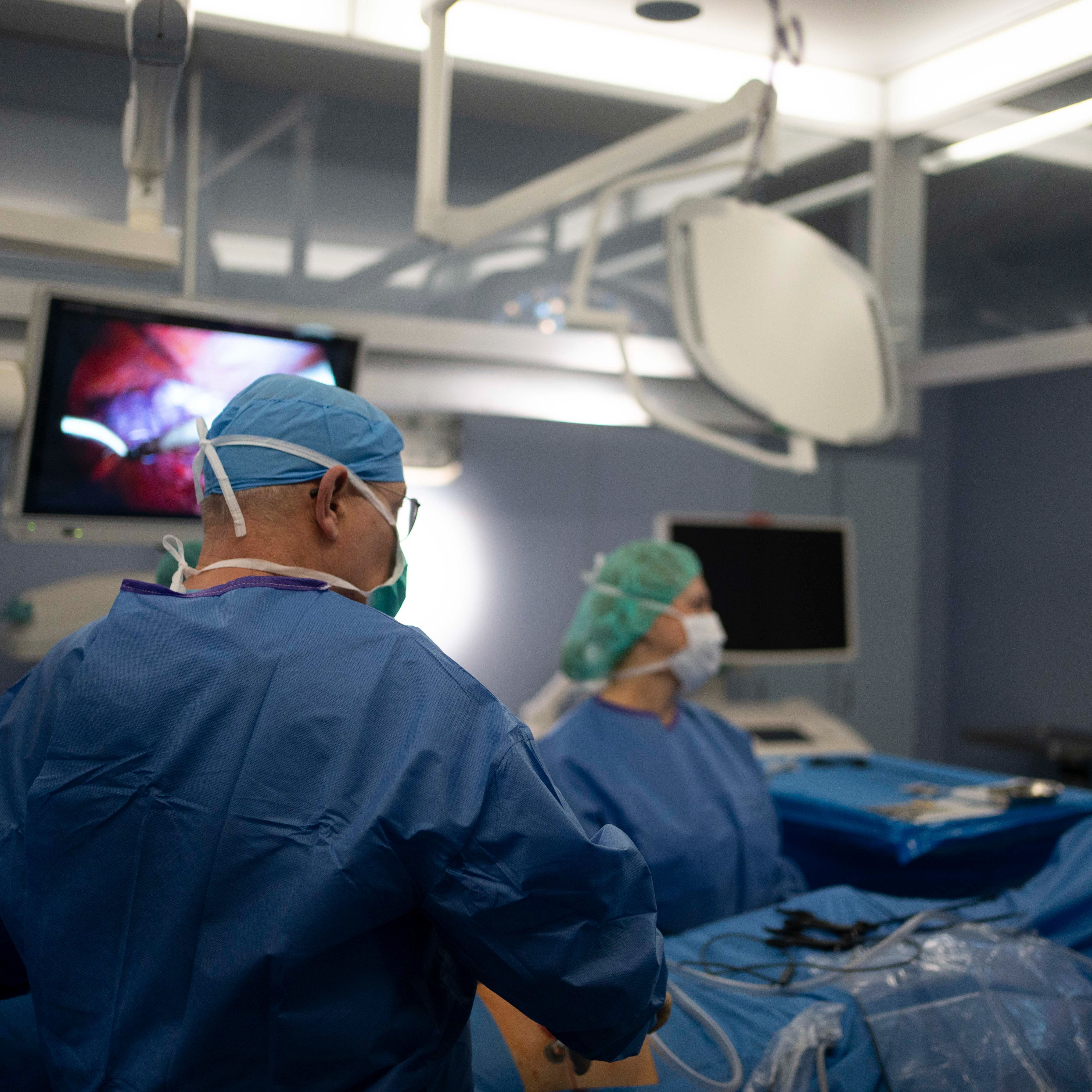 Mehrere medizinische Fachkräfte stehen um ein Krankenhausbett, in dem ein älterer Mann liegt. Sie wirken aufmerksam und besorgt, während der Patient mit geschlossenen Augen ruht.