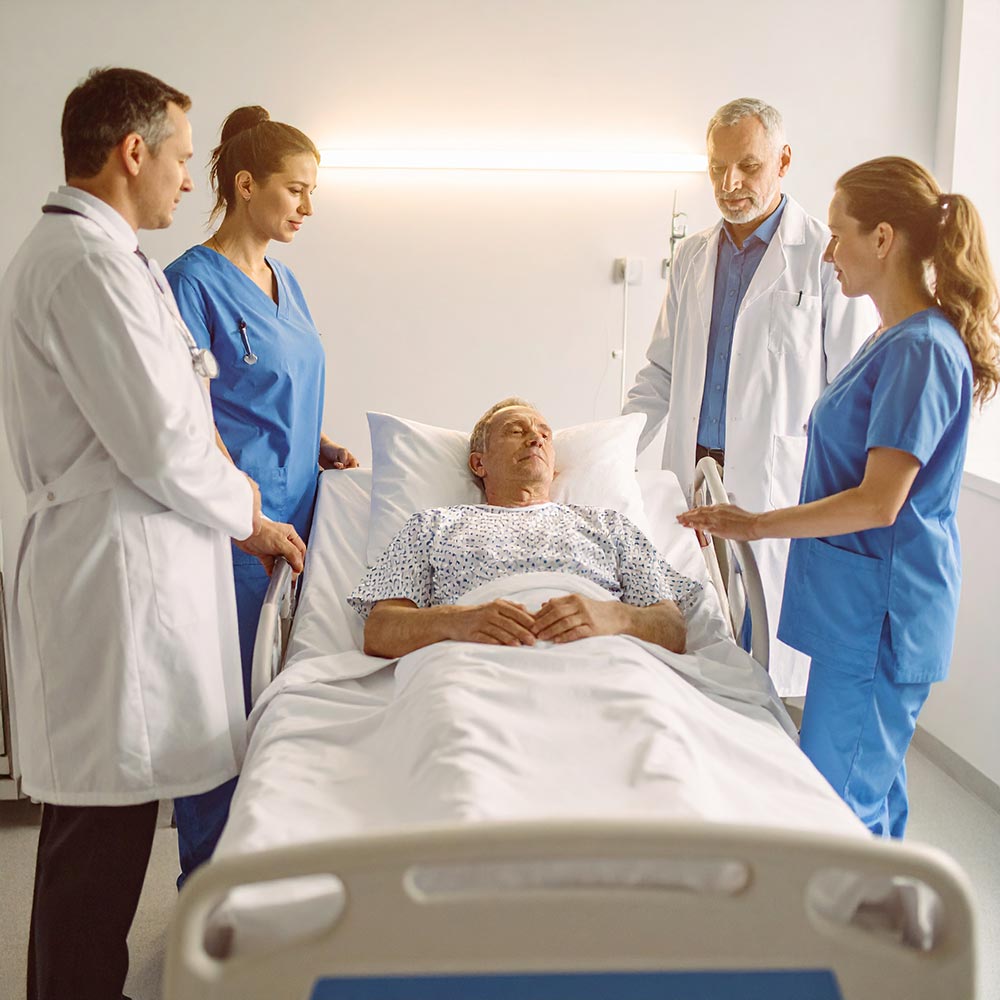 Mehrere medizinische Fachkräfte stehen um ein Krankenhausbett, in dem ein älterer Mann liegt. Sie wirken aufmerksam und besorgt, während der Patient mit geschlossenen Augen ruht.