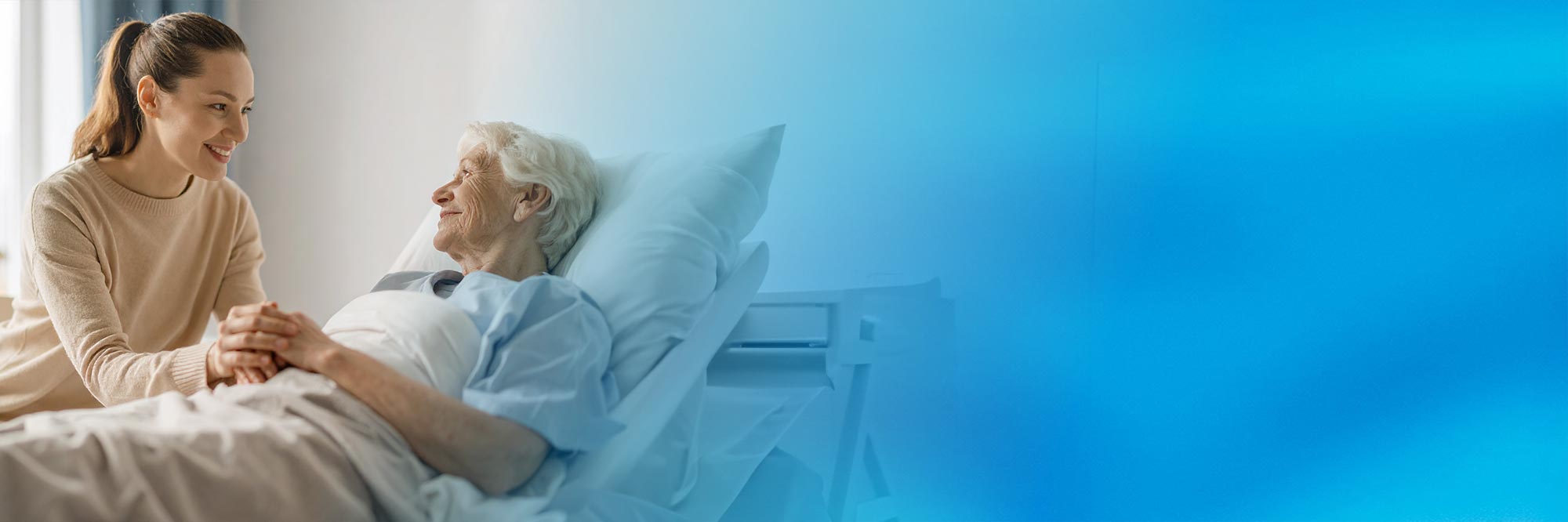 Eine lächelnde ehrenamtliche Besucherin sitzt am Bett einer älteren Patientin, hält ihre Hand und unterhält sich angeregt mit ihr. Der rechte Bildbereich verläuft in einen blauen Farbton.