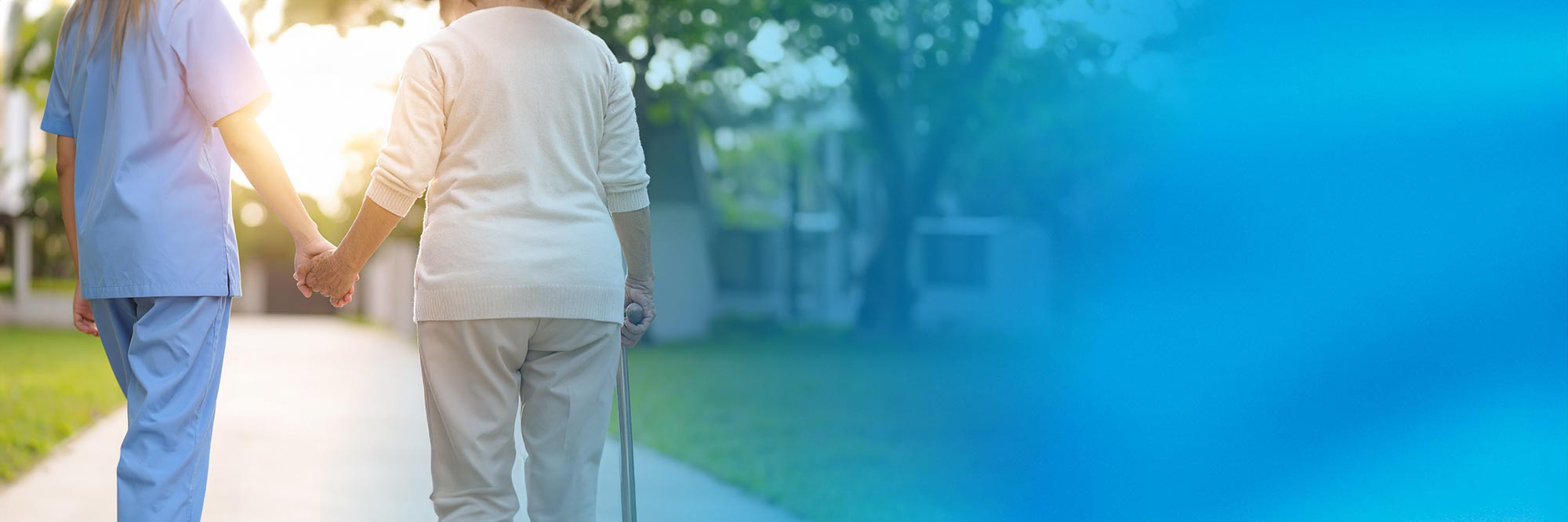 Rückenansicht einer Pflegekraft in blauer Kleidung, die Hand in Hand mit einer älteren Patientin mit Gehstock einen sonnigen Weg im Freien entlanggeht. Der rechte Bildbereich geht in eine blaue Farbfläche über.