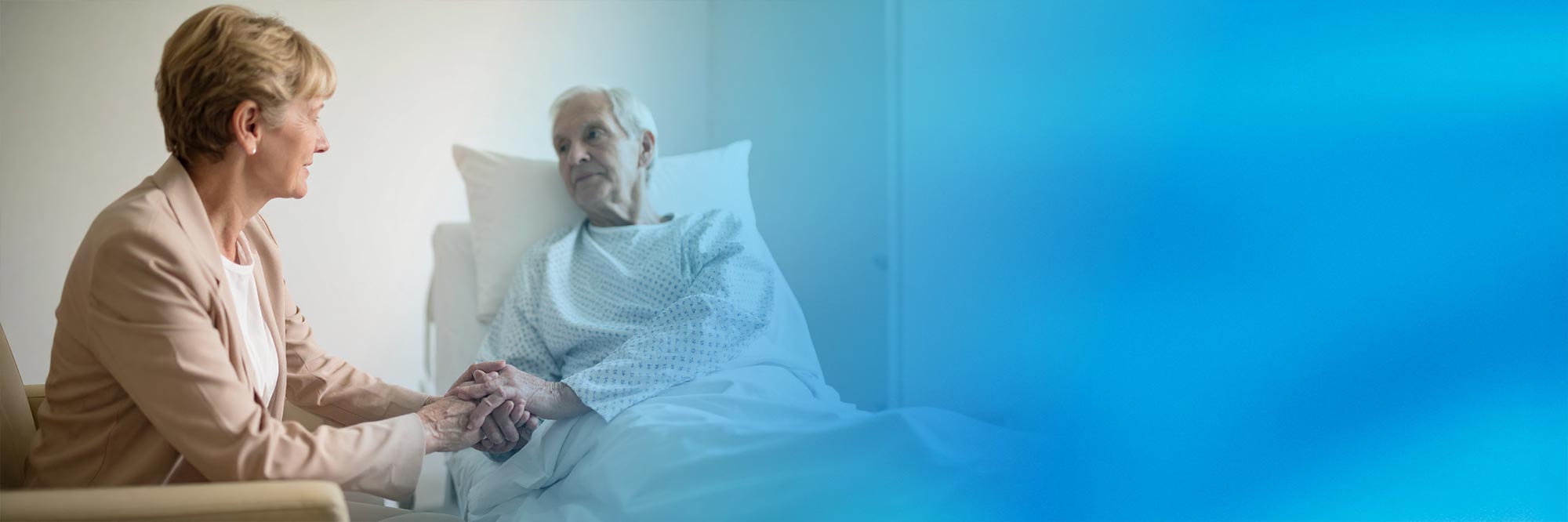 Eine Seelsorgerin in beiger Kleidung sitzt am Bett eines älteren Patienten, hält behutsam seine Hand und sieht ihn zugewandt an. Der rechte Bildbereich geht in einen blauen Farbverlauf über.