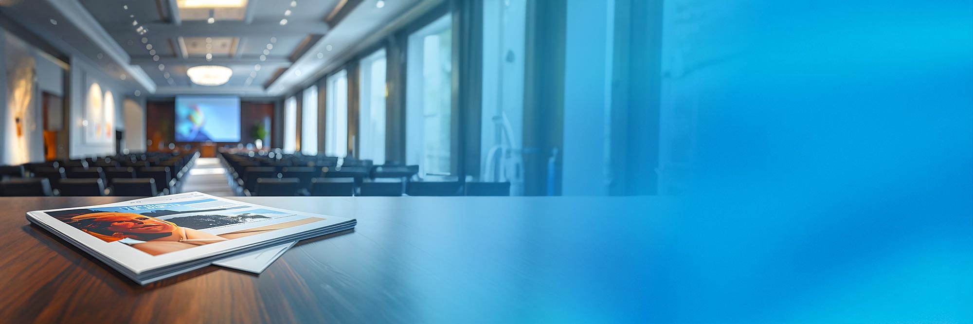 Moderner Konferenzraum mit einem Tisch, auf dem Broschüren liegen. Der Raum ist gut beleuchtet mit natürlichen Lichtquellen aus großen Fenstern, während die Stühle im Hintergrund leer sind. Der Raum wirkt vorbereitet für ein Meeting oder eine Präsentation.