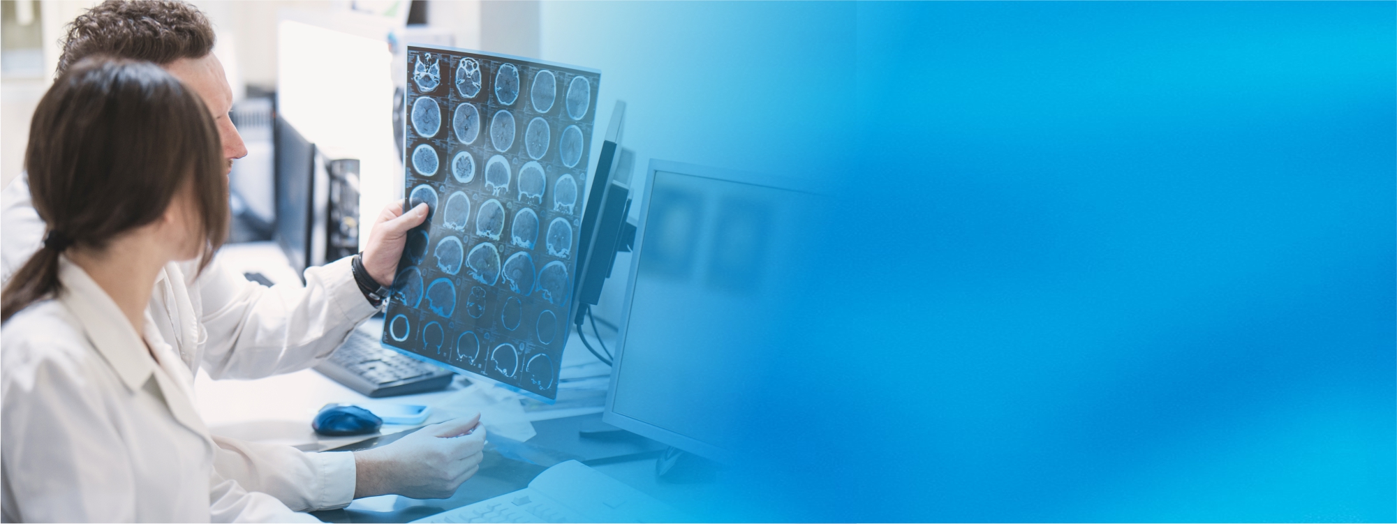 Eine Ärztin und ein Arzt in weißen Kitteln betrachten an einem Computerarbeitsplatz in einer radiologischen Praxis konzentriert den Ausdruck eines Gehirn-Scans. Die rechte Bildhälfte verblasst zu einem blauen Hintergrund.