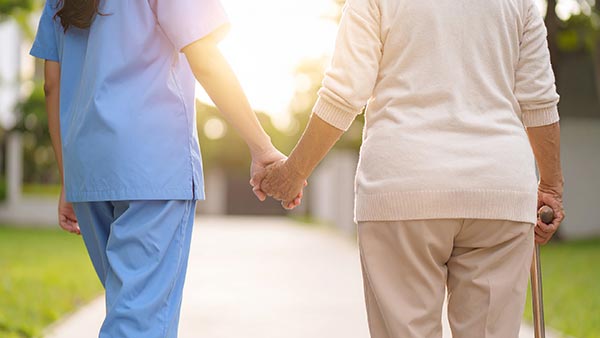 Rückansicht einer Pflegekraft in blauer Berufskleidung, die Hand in Hand mit einer älteren Seniorin mit Gehstock einen sonnigen Weg im Grünen entlanggeht.